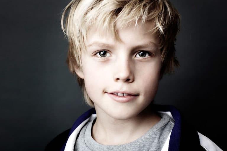 Nærbillede af studieportræt der viser en ung blond dreng, der smiler genert med intense øjne, mod en mørk baggrund.
