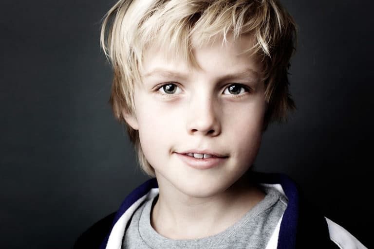 Nærbillede af studieportræt der viser en ung blond dreng, der smiler genert med intense øjne, mod en mørk baggrund.