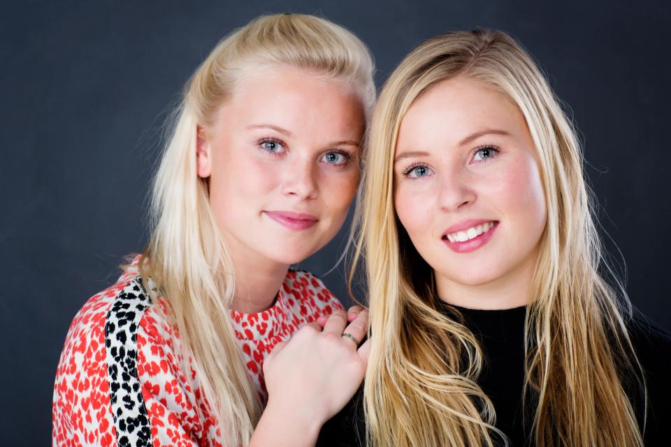 Familiefoto af to smilende blonde søstre med blåt blik, tæt sammen i et autentisk portræt med mørk baggrund.
