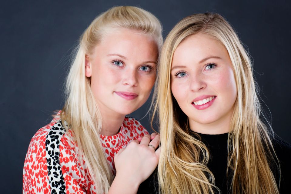 Familiefoto af to smilende blonde søstre med blåt blik, tæt sammen i et autentisk portræt med mørk baggrund.