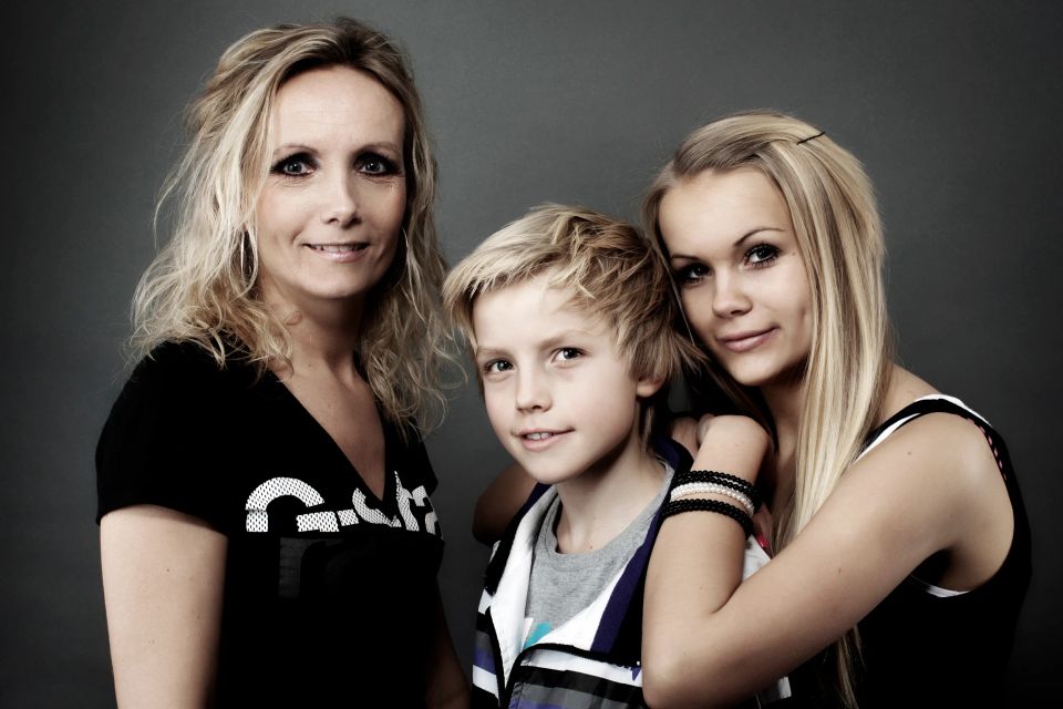 Familieportræt af to blonde kvinder og en dreng med lyst hår, der smiler roligt mod kameraet.