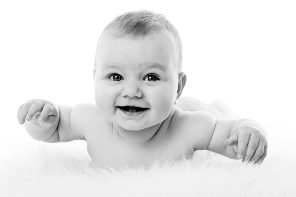 Glad baby ligger på maven på hvidt tæppe og smiler strålende med åben mund i naturligt lys. Babyfotografering.