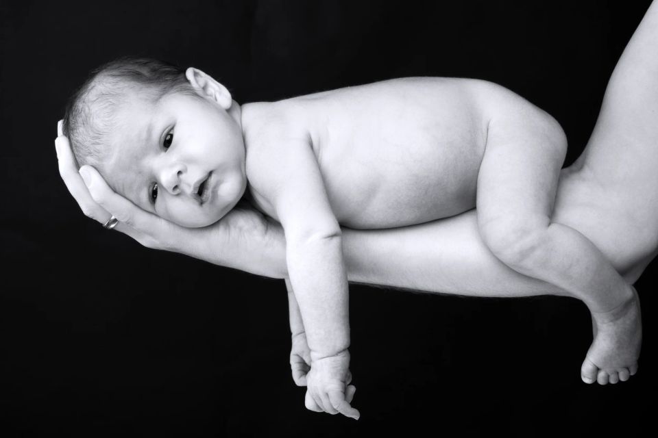 Nyfødt fotografering hvor en baby hviler trygt og nøgen i voksne hænder. S/H, fredfyldt stemning med blødt lys.