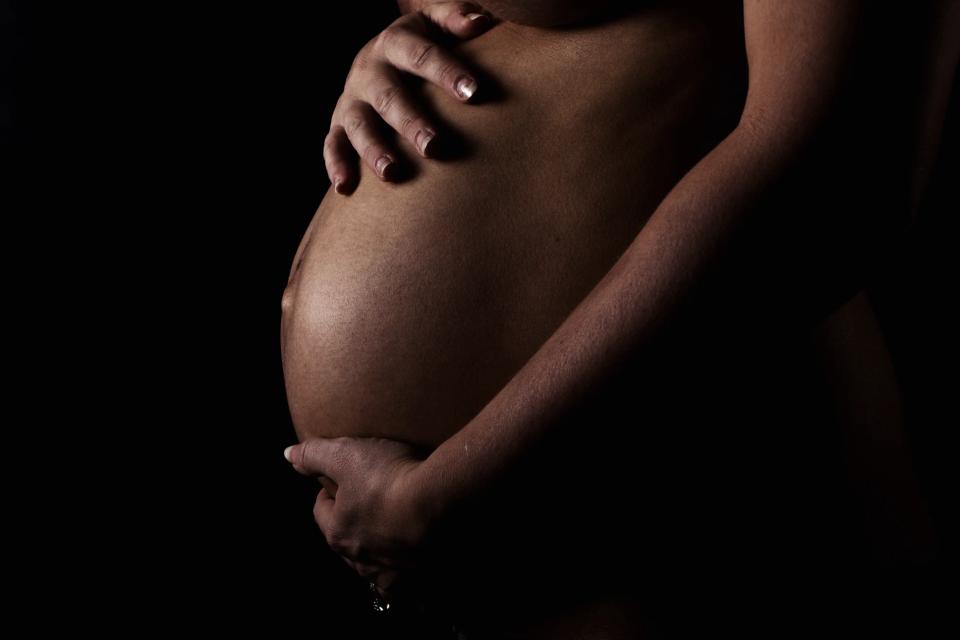 Gravid kvinde omfavner sin store gravide mave i mørket med blødt lys, der fremhæver formen. Fredfyldt gravidfotografering.