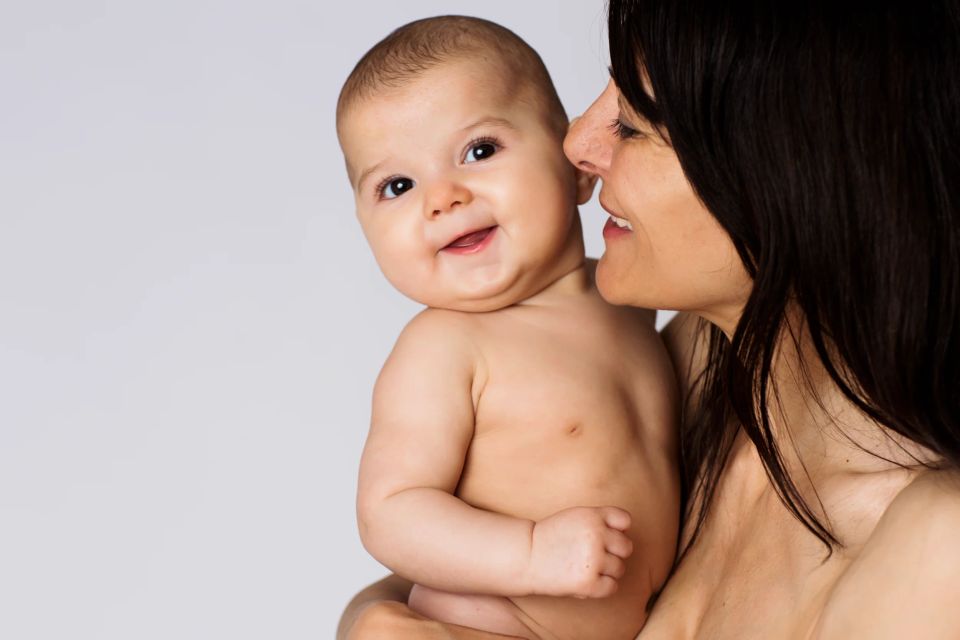 Glad baby fotografering med mor der holder og smiler. Barnet kigger med smil mod kameraet på lys baggrund.