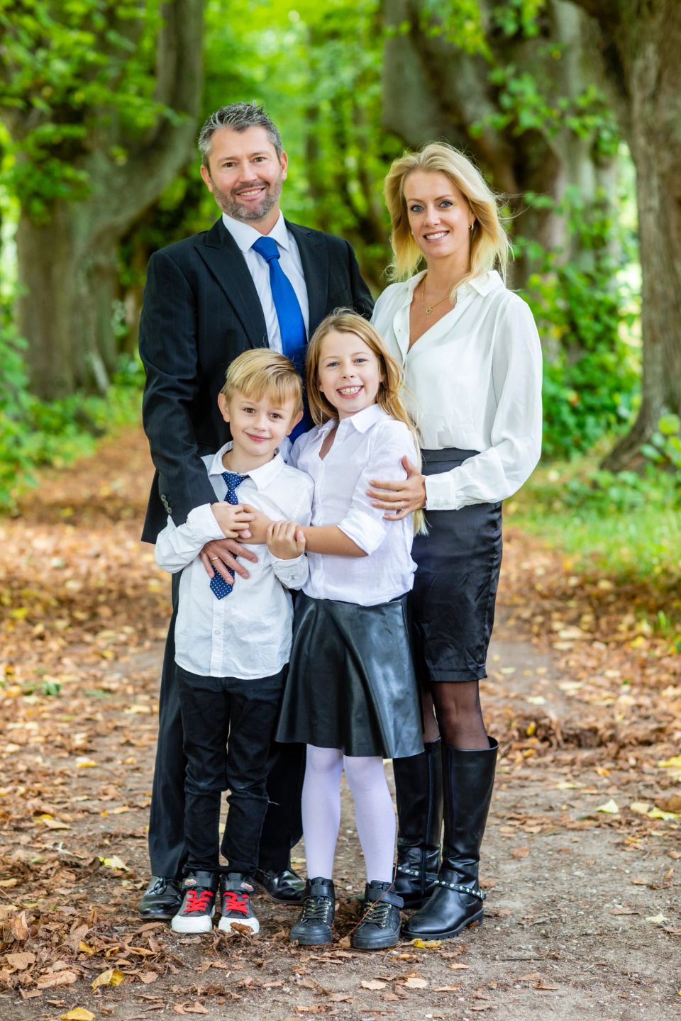 Familiefoto af smilende mor, far og deres to børn klædt i skjorter og festtøj, stående i Virum skov med grønne træer og gy...