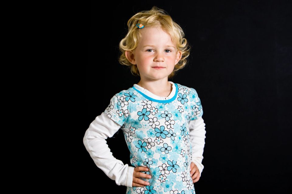 Portrætfoto af et blondt barn med krøller i en blå- og hvidblomstret trøje, der står smilende foran en sort baggrund.