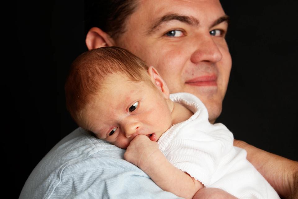 Babyfotografering af en nyfødt, der hviler på sin fars skulder, med faderen smilende blidt mod kameraet.