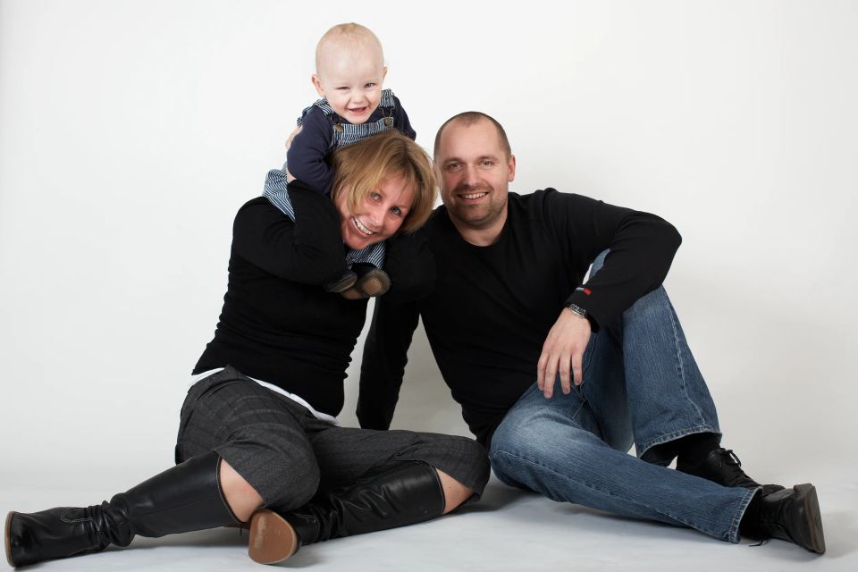 Familieportræt i sort/hvid med forældre siddende og deres glade 1-årige søn på mors skuldre.