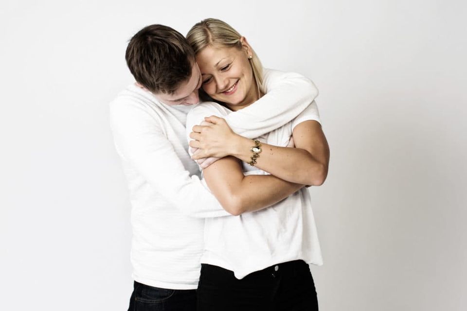 Intimt portrætfoto af kærestepar i hvide bluser der omfavner hinanden med ømme smil, neutral baggrund.