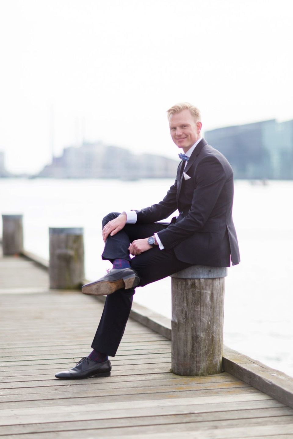 Glad gom i sort jakkesæt med lilla butterfly sidder på pæl ved Islands Brygge kajkant. Bryllupsfotografering.