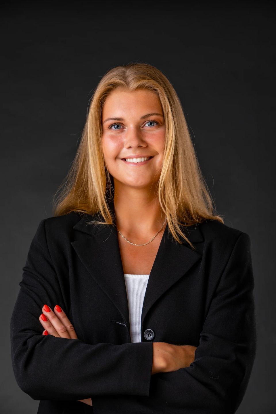 Portrætfoto af ung, smilende kvinde med blondt hår, hvid top og sort blazer foran en mørk baggrund.