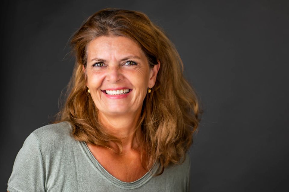 Glad kvinde med smilende ansigt i autentisk portrætfoto. Lyst hår blæser let, baggrund er mørk og neutral.