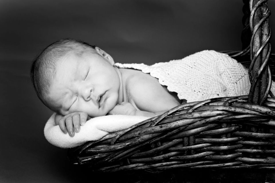 Sort-hvid babyfotografering af en nyfødt sovende trygt i en flettet kurv med blødt tæppe.