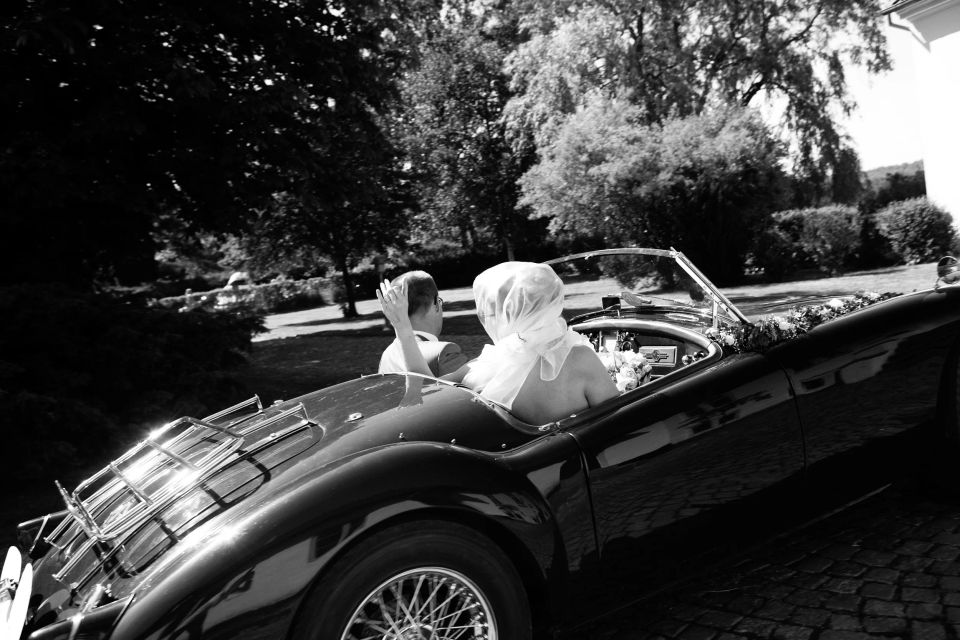 Sort-hvidt bryllupsfoto af brudepar i sort, åben sportsvogn på brosten, gæster vinker i baggrunden i solskin.