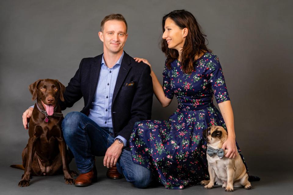 Portrætfoto af par og to hunde: en labrador og en mops foran grå baggrund. Kvinden kigger kærligt på manden.
