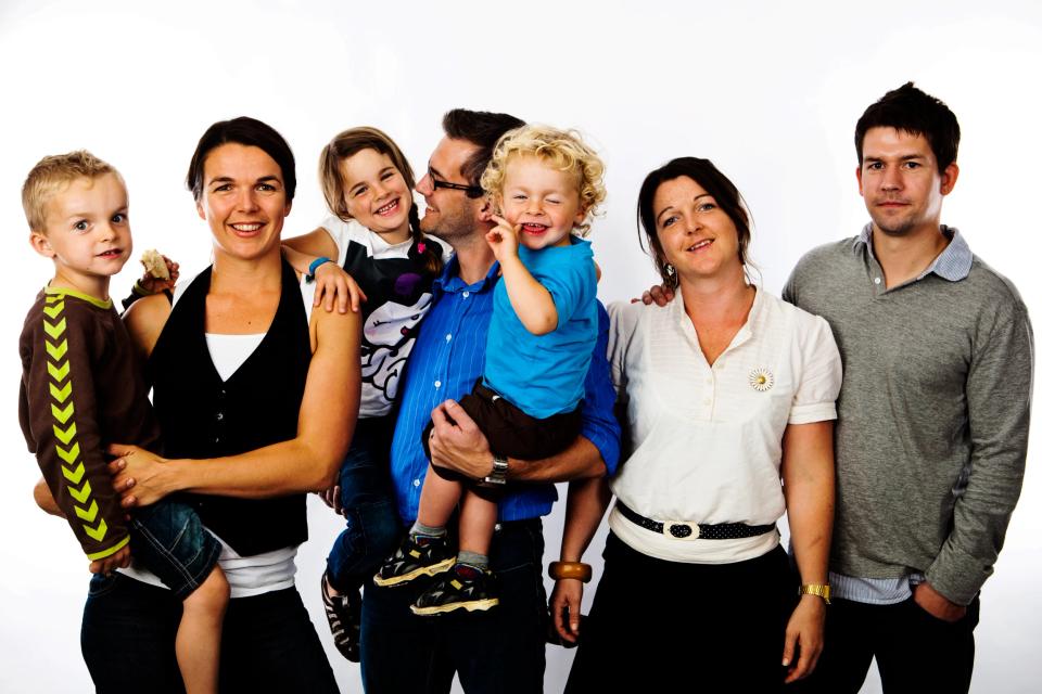Autentisk familiefoto af gruppe på seks glade individer, tre voksne og tre legende børn i hvid studiebaggrund.