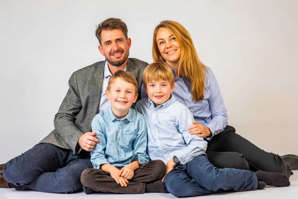 Familiefoto af smilende mor, far og to drenge siddende tæt sammen mod en lys baggrund. Blødt studie fotografi.