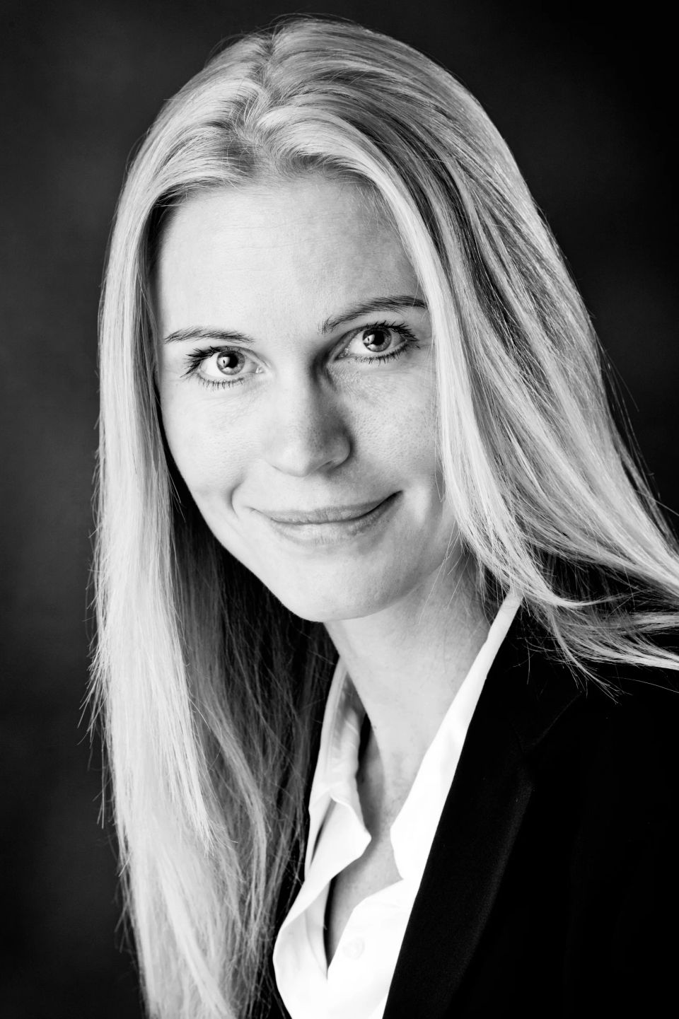 Portrætfoto af blond kvinde i sort blazer med hvid skjorte, der smiler imødekommende med et professionelt udtryk.