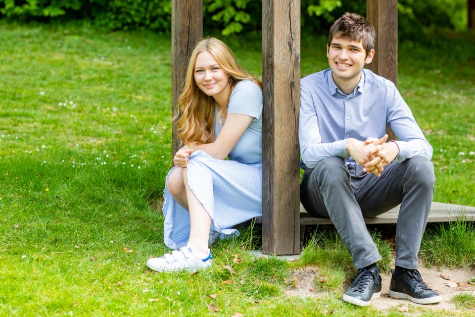 Søskendefoto af ung kvinde og mand smilende siddende i græsset mellem træbjælker i naturligt lys.