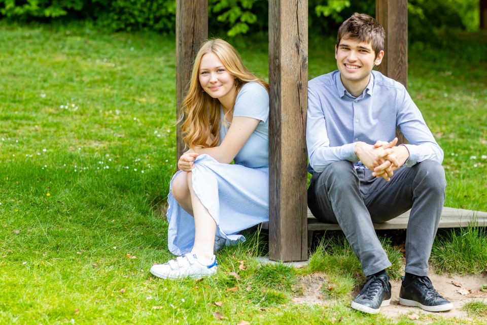 Søskendefoto af ung kvinde og mand smilende siddende i græsset mellem træbjælker i naturligt lys.