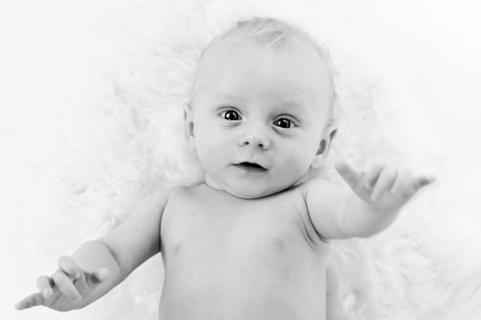 Sort/hvidt babyportræt af nysgerrig baby liggende på ryggen med store øjne og udstrakte arme på et blødt underlag.