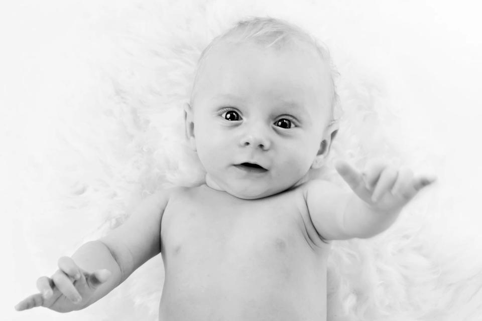 Sort/hvidt babyportræt af nysgerrig baby liggende på ryggen med store øjne og udstrakte arme på et blødt underlag.