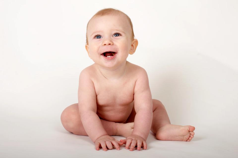 Glad baby griner i charmerende portrætfoto. En nøgen baby sidder og smiler bredt med åben mund og viser to tænder.