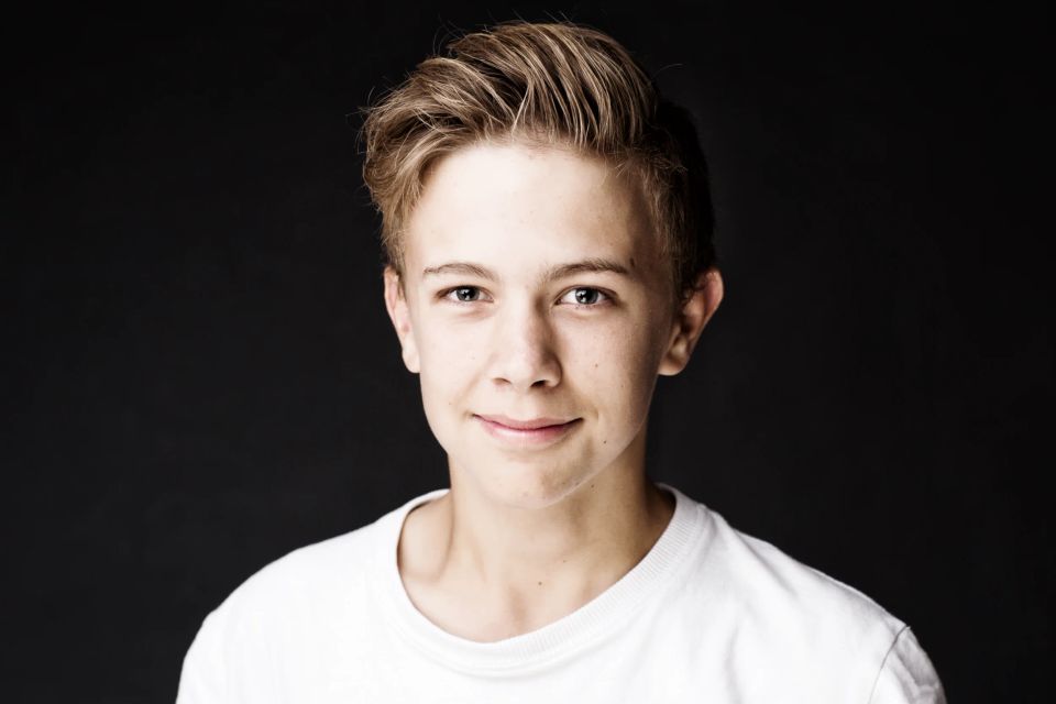 Nært portrætfoto af ung dreng med lyst hår og charmerende smil mod mørk baggrund i naturligt lys. Børnefotografering.