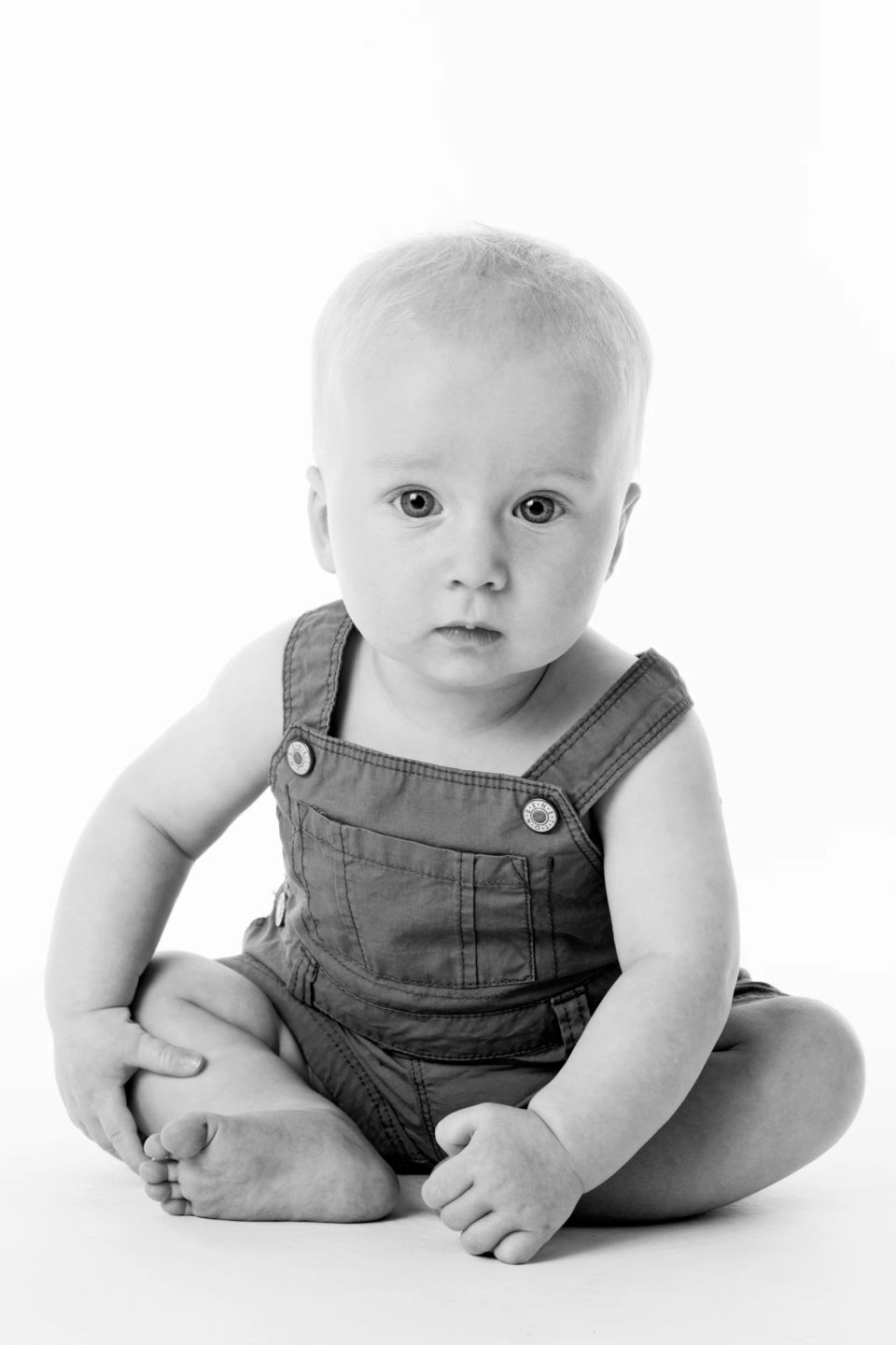 Babyfotografering af siddende lyshåret baby i blå selebukser med et nysgerrigt barnedåbsblik mod beskueren.