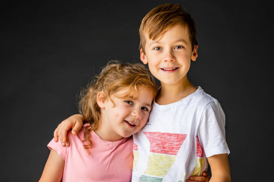 Familiefoto af søskende: En smilende dreng med armen om sin lillesøster, der læner hovedet mod ham, mod mørk baggrund.