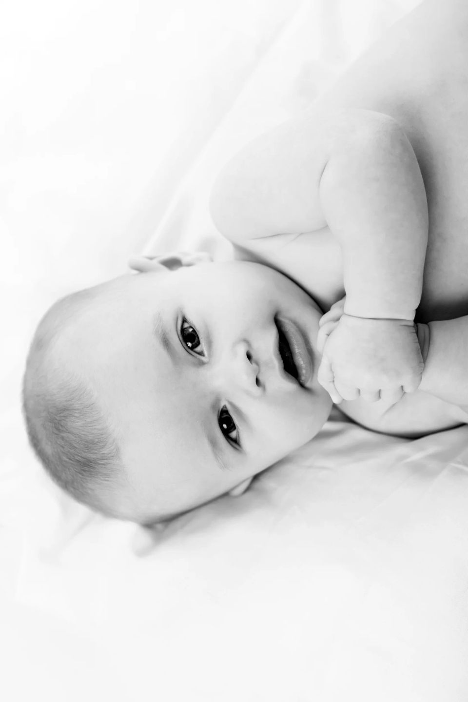 Sort/hvid babyportræt af ægte, nuttet baby liggende på maven med hænderne samlet. Blød belysning og hvid baggrund.