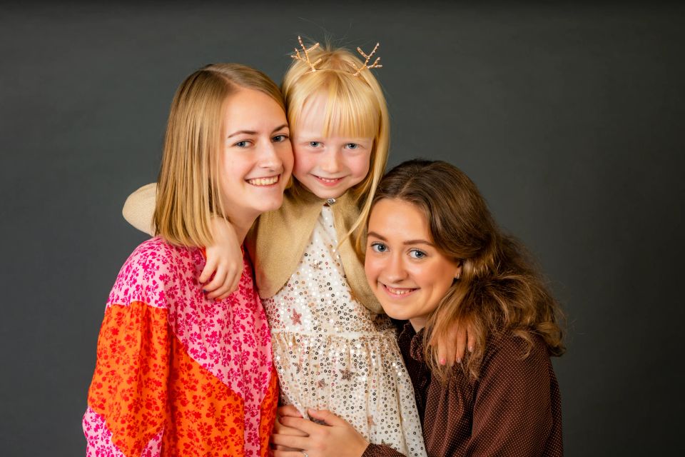 Familiefotografi af tre smilende søstre, hvoraf den yngste bærer et diadem, der omfavner hinanden rørende.