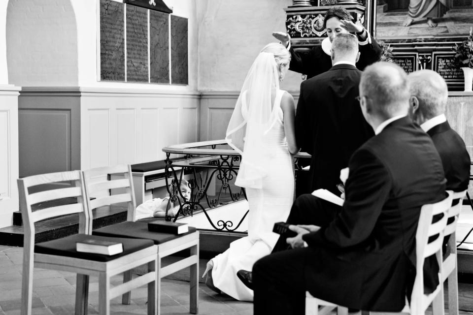 Bryllupsbillede: Et nysgerrigt barn kigger frem under stole ved vielsen i kirken, sort/hvidt foto.