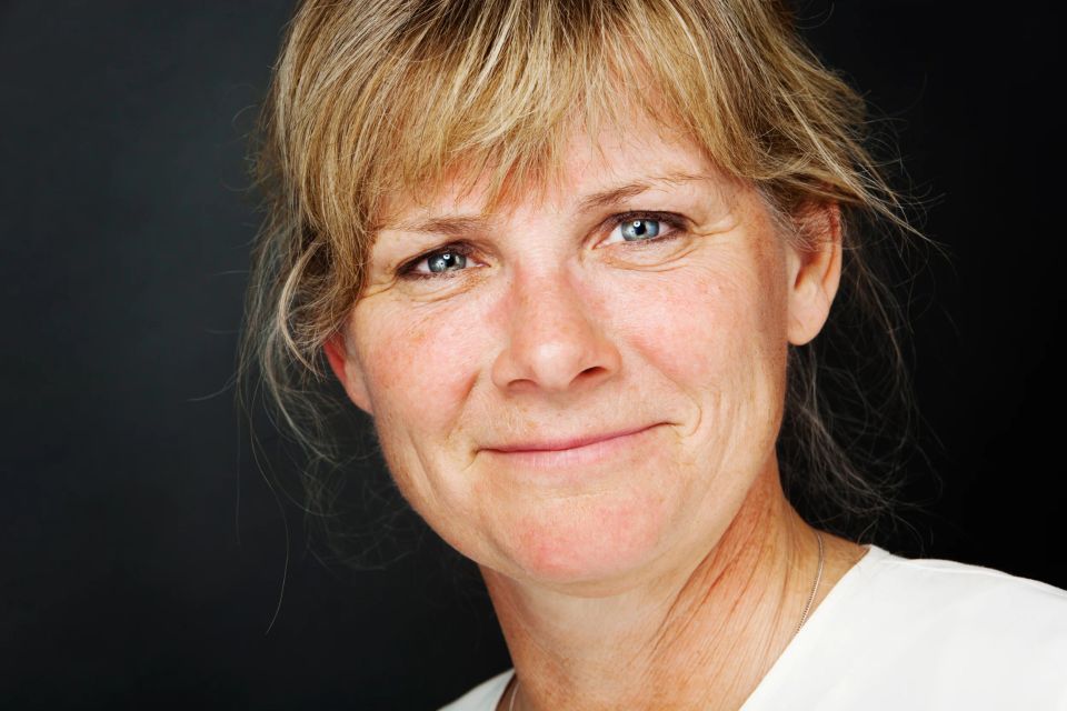 Nært portrætfoto af smilende kvinde med lyseblå øjne og blondt hår. Blød belysning mod mørk baggrund, skaber en varm stemn...