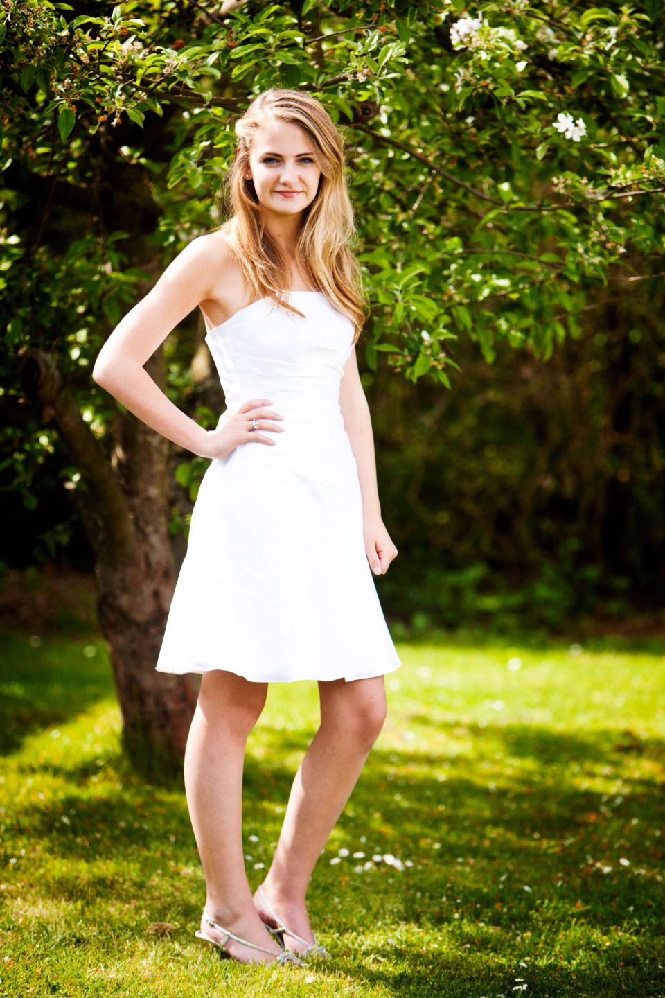 Portrætfoto af ung kvinde i hvid stropløs kjole, stående i grøn have med sollys og træ i baggrunden.
