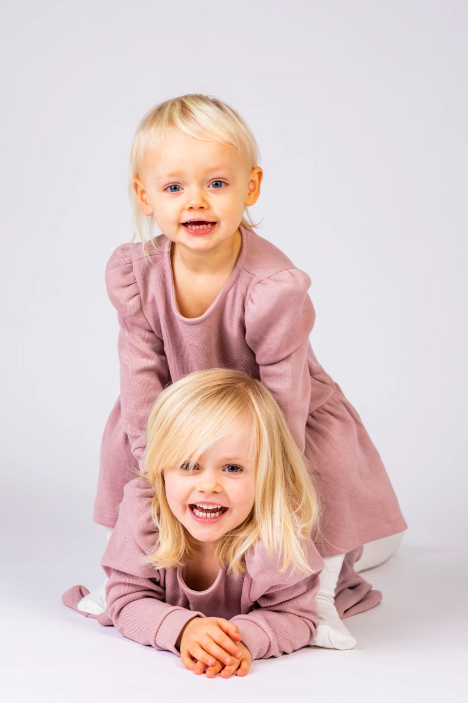 To legende blonde børn i rosa tøj smiler stort mod kameraet under en fotografering med hvid baggrund.