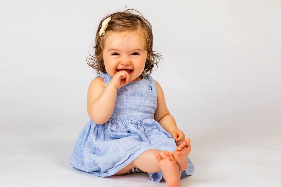 Børnefotografering: Glad, grinefuld baby i lyseblå kjole og hårspænde sidder på gulvet. Lyse omgivelser. Virum