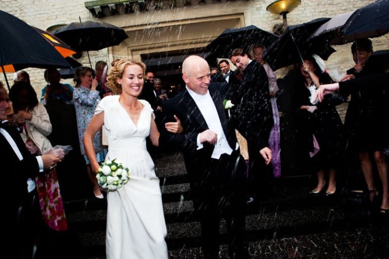 Bryllupsfotografering af smilende brudepar i regnvejr med paraplyer holdt af gæster, der kaster ris.