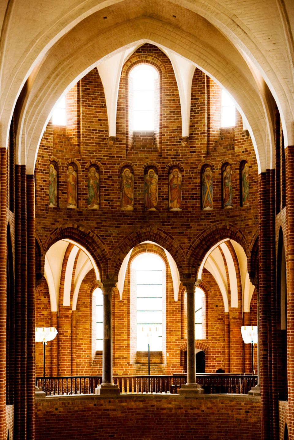 Historisk kirkeinteriør med røde mursten, buer, lyse vinduer og udskårne helgenfigurer, stemningsfuld stemning.