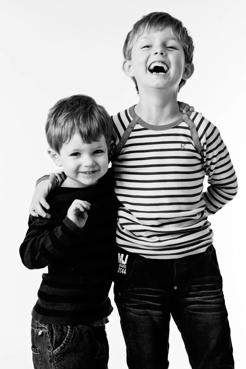 To legesyge drengebørn i sort/hvid stribet tøj, griner og smiler tæt sammen foran hvid baggrund i et professionelt børnefoto.