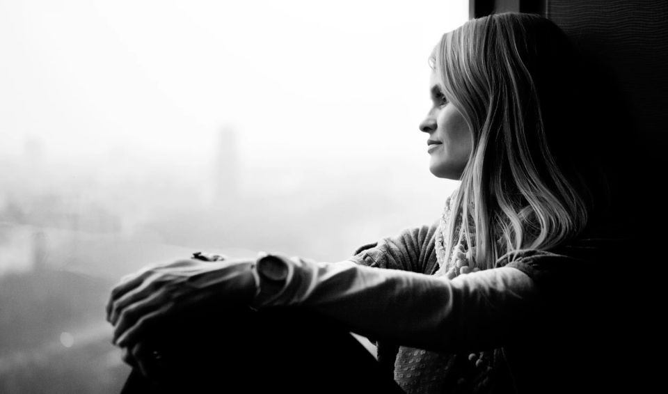Sort/hvidt portrætbillede af en kvinde med lyst hår, der kigger eftertænksomt ud af et vindue med blødt lys.
