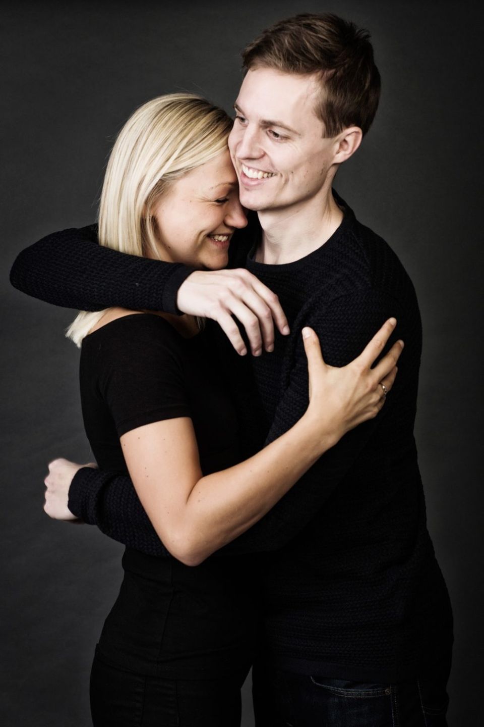 Portrætfoto af forelsket par der krammer tæt med glæde og smil, mørk baggrund, blødt lys.