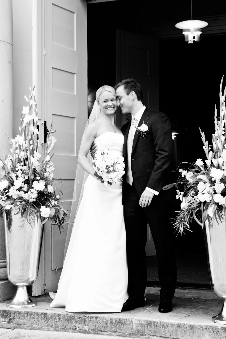 Bryllupsfotografering: Smilende brudepar i omfavnelse foran kirken i Virum, omgivet af hvide blomsterarrangementer.