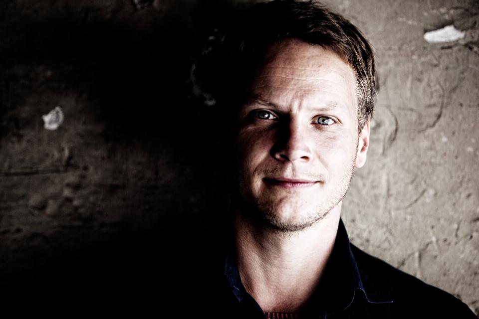 Portrætfoto af ung mand med blåt øje og mørkt hår, delvist belyst fra venstre mod en grov væg i dæmpet belysning.