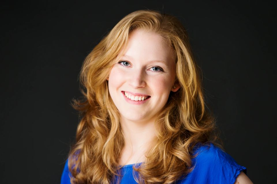 Portrætfoto af smilende kvinde med langt, lyst hår og blå bluse mod en mørk baggrund. Oprigtig glæde fotograferet.