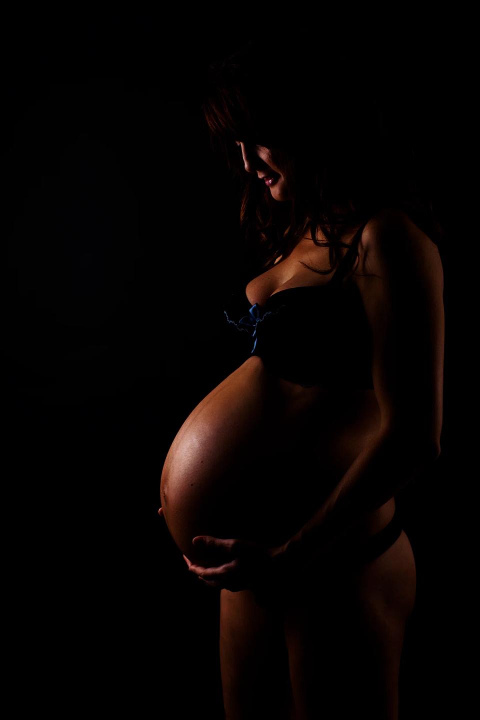 Gravid kvinde i profil med hænderne på sin mave i et stemningsfuldt graviditetsportræt med blødt lys.