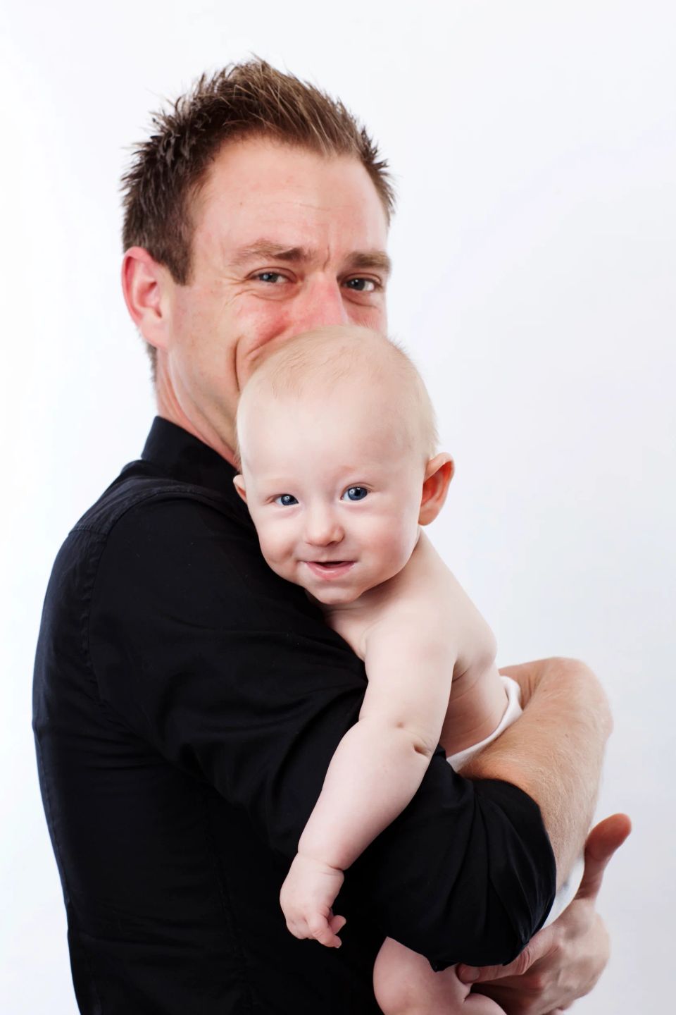 Babyfotografi af smilende far, der holder sin glade baby. Babyen kigger frem med blå øjne i blødt lys.