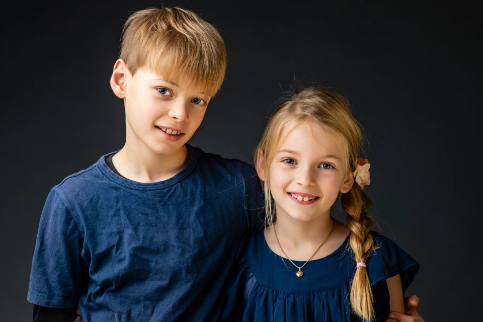 Familiefotografering med to søskende, der smiler varmt til kameraet, den ene arm om den anden. Naturligt lys.