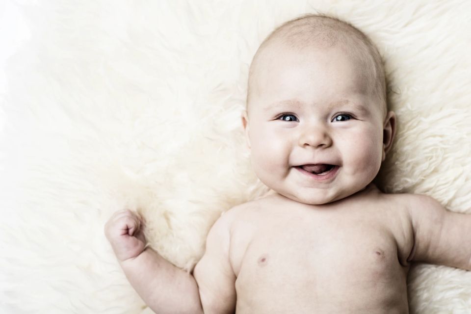 Babyfotografering: Smilende glad baby ligger på blødt, lyst tæppe og ser direkte mod kameraet med åben mund.